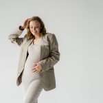 Cheerful pregnant woman in dress and jacket posing isolated on grey